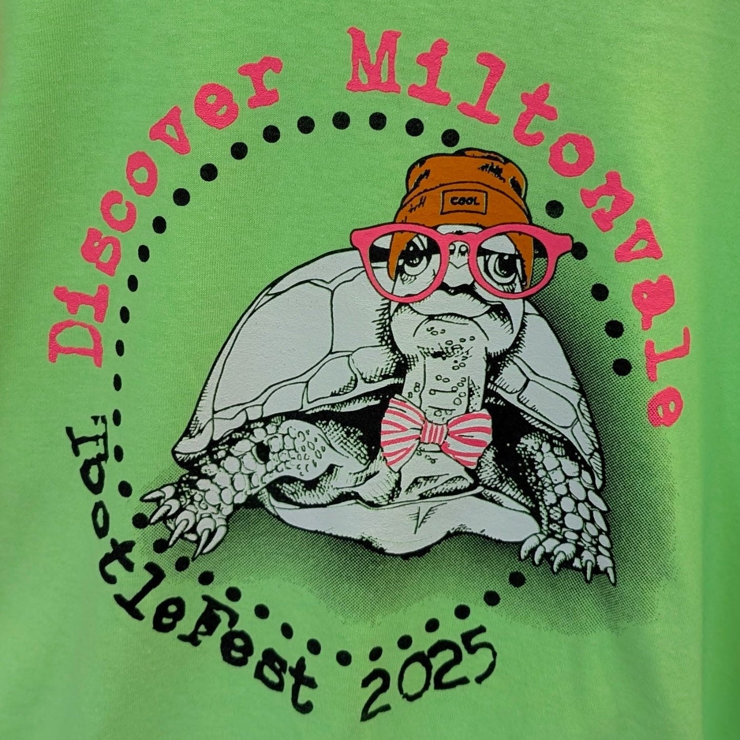 Green t-shirt with a graphic of a tortoise wearing glasses and a bow tie, and text 'Discover Miltonvale TurtleFest 2025'.