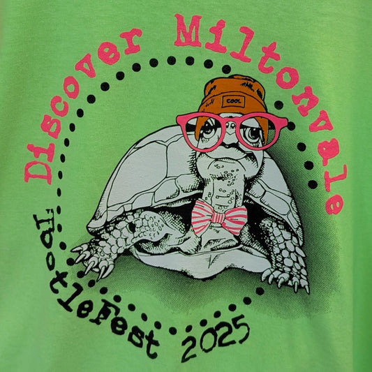 Green t-shirt with a graphic of a tortoise wearing glasses and a bow tie, and text 'Discover Miltonvale TurtleFest 2025'.